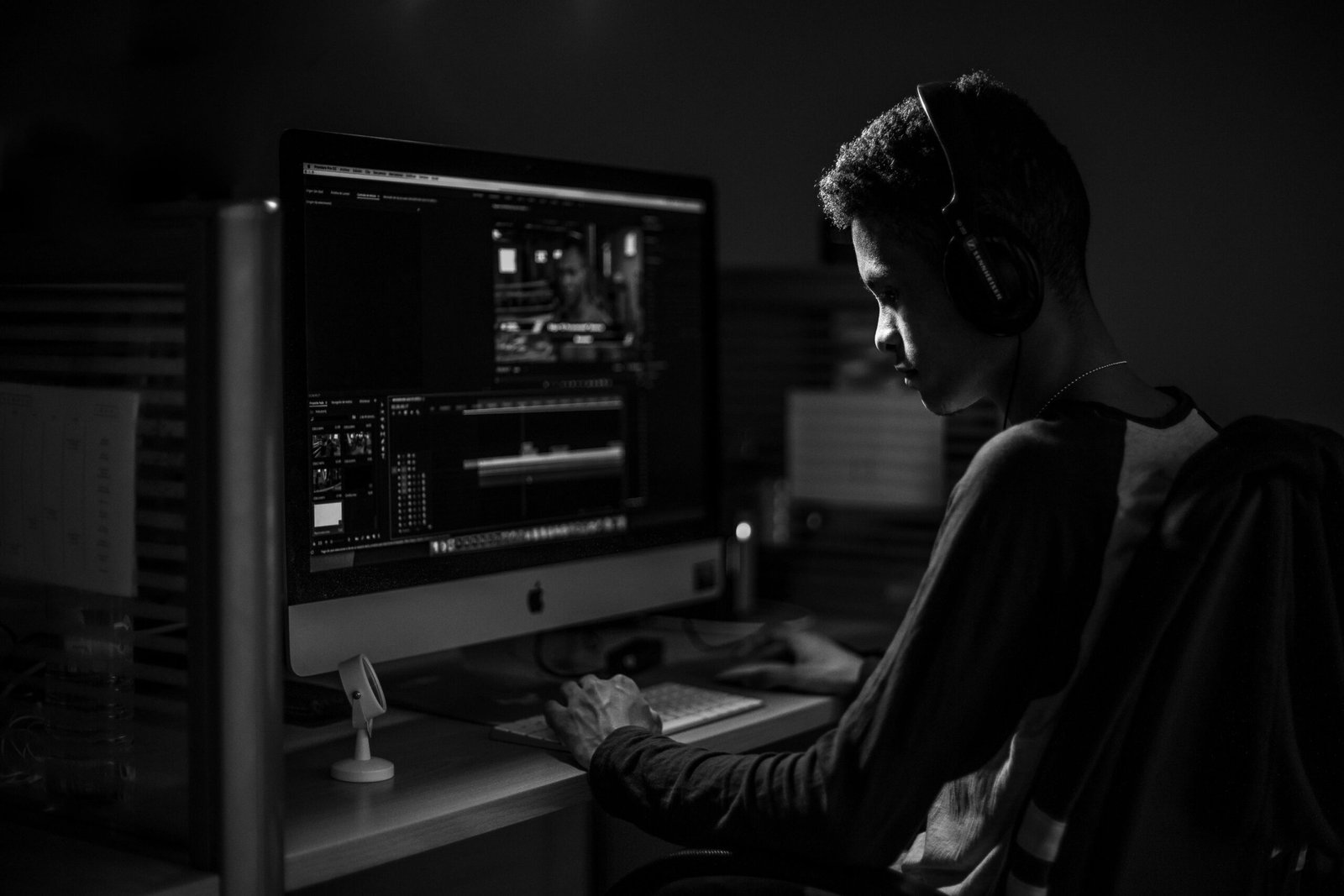 A man wearing headphones working on video editing on a computer in a dimly lit office.